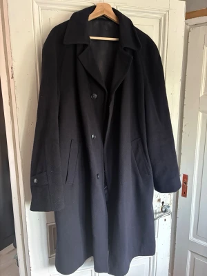 ORATOP herrock, Ull & 30% Kashmir  - Stilren svart ullrock med knäppning framtill och två sidofickor. Rocken har bred krage och knappar även vid ärmsluten. Perfekt för dig som vill ha en tidlös och enkel look under kallare dagar.