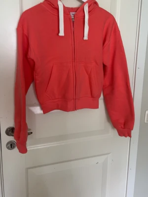Korallfärgad hoodie från Gina Tricot - Säljer en snygg korallfärgad hoodie från Gina Tricot i storlek XS. Hoodien har dragkedja framtill, vita snören i huvan och en stor magficka. Tillverkad i mjukt sweatshirtmaterial och har ribbade muddar vid ärmar och nederkant.