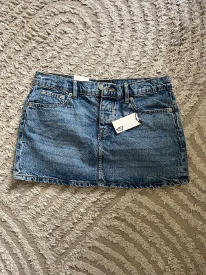 Blå jeanskjol i kort modell - Säljer en klassisk blå jeanskjol i kort modell med fem fickor och bälteshällor. Kjolen har en rak passform och stängs med knapp framtill. Den är aldrig använd! 💕