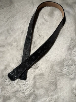 Lv bälte - Säljer ett svart Louis Vuitton-inspirerat bälte med Monogram Eclipse-mönster och svart LV-spänne. Stilren modell som passar till både vardags- och festoutfits. Bältet är i använt skick med synligt slitage vid några av hålen samt normalt bruksslitage, se bilder för egen bedömning.