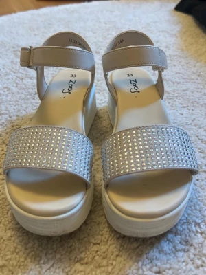 Beige platåsandaler med strass - Snygga beige platåsandaler med breda remmar täckta av glittrande strass och justerbart spänne runt ankeln. Sandalerna har en hög, vit platåsula och är tillverkade i syntetmaterial. Perfekta för dig som vill sticka ut lite extra.