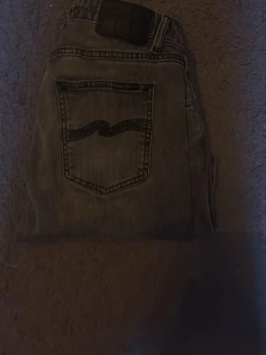 Nudie jeans w28 - Säljer ett par grå jeans med rak passform och klassisk femficksdesign. Jeansen har en tydlig vågig söm på bakfickan och en mörkare tvätt för en cool vintagekänsla. Perfekta för dig som gillar avslappnad stil och tidlös denim. Priset är inte hugget i sten hör av er vid minsta fundering.