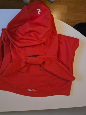 Orange hoodie från Peak Performance - Säljer en snygg orange hoodie från Peak Performance med vit logga på bröstet och en liten logga på huvan. Hoodien har klassisk passform, stor huva och ribbade muddar. Perfekt för dig som vill ha en stilren och bekväm look.