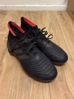 Adidas predator  - Snygga svarta fotbollsskor med högt skaft och röda detaljer på insidan. Skorna har snörning och mönstrad ovansida för bättre bollkontroll. Utrustade med fasta dobbar för spel på gräs. Tillverkade i syntetmaterial för bra passform och hållbarhet.