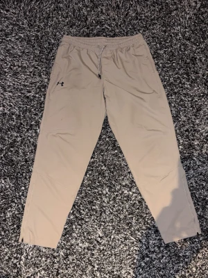  Under Armour - Snygga beige mjukisbyxor från Under Armour med dragsko i midjan och raka ben. Byxorna har diskreta fickor och dragkedja vid benslutet för extra stil och funktion. Perfekta för chill eller träning.
