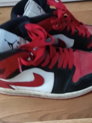 Nike Air Jordan 1 svart/röd/vit - Säljer ett par Nike Air Jordan 1 sneakers i svart, rött och vitt. Skorna har röda snören, vit tunga med svart Jumpman-logga och klassisk röd swoosh på sidan. Ovandelen är i läder och designen är ikonisk med högt skaft.