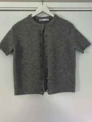Grå kortärmad kofta från Zara - Kortärmad grå kofta från Zara med rund halsringning och ribbade kanter. Koftan har knappar framtill i metall och är gjord i ett mjukt stickat material. Perfekt att styla över en topp eller klänning för en enkel och clean look.