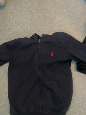 Mörkblå hoodie från Polo Ralph Lauren - Snygg mörkblå hoodie med dragkedja från Polo Ralph Lauren. Klassisk röd logga broderad på bröstet och två fickor framtill. Perfekt för en chill och stilren look. Mjuk bomullskänsla och ribbade muddar vid ärmslut och nederkant. Den är helt ny har använt den bara 3 gånger inga defekter fel eller sak på storleken är m men passar för bägge m och s