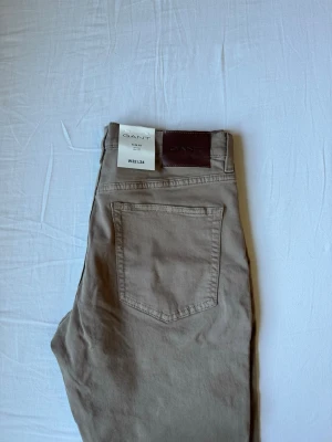 Beige jeans från GANT - Snygga beige jeans från GANT med klassisk femficksmodell och raka ben. Jeansen har en stilren look med läderpatch bak i midjan och metallknappar. Tillverkade i mjukt bomullsmaterial som ger en skön känsla och passar perfekt till vardagslooken.