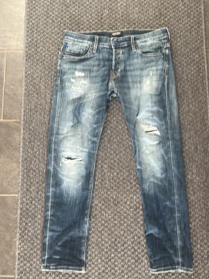 Blå slimfit jeans från jackandjones - Säljer ett par blå jeans med slitningar och hål på knäna. Jeansen har en klassisk femficksmodell, raka ben och tydliga tvättade detaljer för en cool, avslappnad look. Perfekta för dig som gillar streetstyle och en trendig vibe.