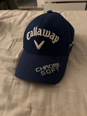 Blå Callaway keps med brodyr - Snygg blå keps från Callaway med vit brodyr på framsidan där det står 'Callaway' och 'Chrome Soft'. På sidan finns även texten 'Apex'. Klassisk böjd skärm och justerbar passform. Perfekt för golf eller streetwear.