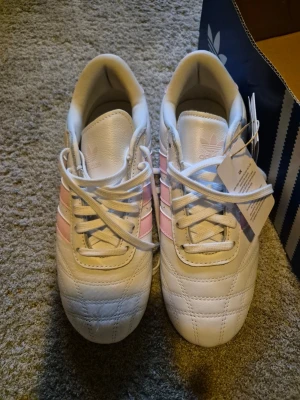 Vita sneakers med rosa ränder Adidas - Snygga vita sneakers från Adidas med klassiska tre rosa ränder på sidan. Skorna har en låg profil, vita snören och ovandel i vad som ser ut som läder. Perfekta för dig som vill ha en clean och sportig look med en touch av färg.