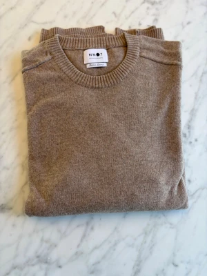 Beige stickad tröja från NN07 - Snygg beige stickad tröja från NN07 med rund halsringning och ribbade muddar vid ärmslut och nederkant. Tröjan har en klassisk och stilren design, perfekt för lager på lager och enkel att matcha med olika outfits. Materialet är mjukt och bekvämt. Oanvänd. Nypris cirka 1400 SEK. 