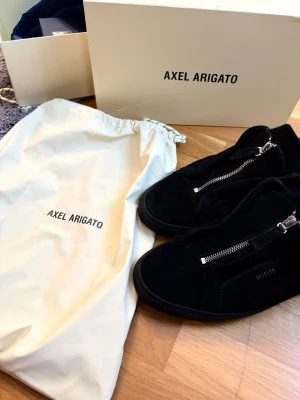 Svarta sneakers från Axel Arigato - Snygga svarta sneakers från Axel Arigato i mocka med silverfärgade dragkedjor på ovansidan. Skorna har en minimalistisk design och låg profil. Perfekta för dig som gillar stilrena och moderna detaljer. Storlek 44-45