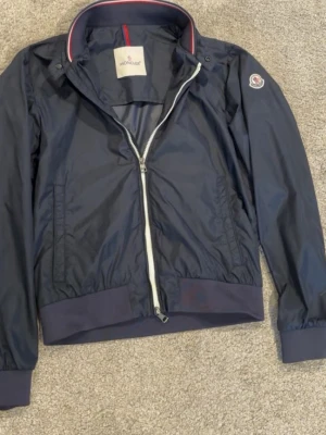 Moncler Windbreaker   - Snygg mörkblå vindjacka från Moncler med vit dragkedja, ribbade muddar och krage med röd och vit detalj. Jackan har Moncler-logga på ärmen och är tillverkad i ett tunt, glansigt material som står emot vind. Perfekt för dig som vill ha en stilren och sportig look.