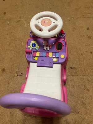 Rosa prinsessbil med ratt och slott - Säljer en söt rosa och lila leksaksbil med prinsesstema. Bilen har ett vitt säte, ratt, knappar med ljud och ljus samt ett slott med små prinsessfigurer på baksidan. Perfekt för yngre barn som gillar prinsessor och fantasifulla lekar.