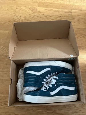 Vans  - Snygga blåa höga sneakers med vit sula och vita detaljer. Skorna har klassisk vågig sidostrip och vita skosnören. Ovandelen är i mockaliknande material och yttersulan är i gummi med rutmönster. Perfekta för dig som gillar streetwear och vill ha en stilren look.