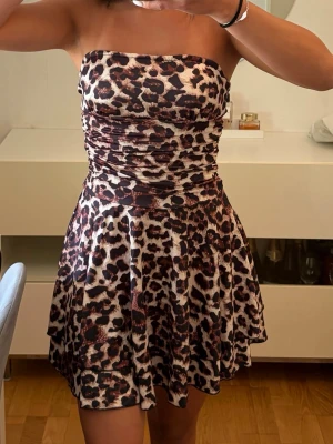 Axelbandslös leopardklänning - Säljer en axelbandslös kort klänning med leopardmönster. Klänningen har en figurnära överdel och utställd kjol, perfekt för dig som vill sticka ut. Materialet känns mjukt och följsamt, troligen polyester. Supersnygg till sommarens alla tillfällen!