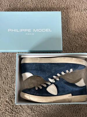 Blå Philippe Model sneakers i mocka - Snygga blå sneakers från Philippe Model i mocka med vita detaljer och vit sula. Skorna har en låg silhuett, vita skosnören och en ikonisk sköldlogga på sidan. Perfekta för dig som gillar stilrena och trendiga sneakers.