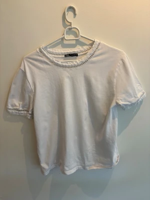 Vit t-shirt med spetsdetaljer - Säljer en vit t-shirt från Zara med korta ärmar och snygga spetsdetaljer vid halsringning och ärmslut. Tillverkad i mjuk bomull och har en klassisk passform som passar till det mesta. 