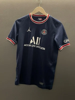 Mörkblå PSG matchtröja - Snygg mörkblå PSG matchtröja med vita och röda detaljer vid krage och ärmslut. Tröjan har klubbmärke, Jordan-logga och sponsortryck på bröstet samt tryck på ena ärmen. Tillverkad i ett lätt och ventilerande material, perfekt för fotboll eller träning.