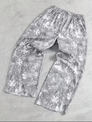 Grå camo mjukisbyxor med lövprint - Säljer ett par grå mjukisbyxor med bred passform och elastisk midja. Byxorna har ett unikt camo-mönster med vita löv och grenar, samt dragsko i midjan för extra komfort. Perfekta för en chill och avslappnad stil. Ny och storlek M
