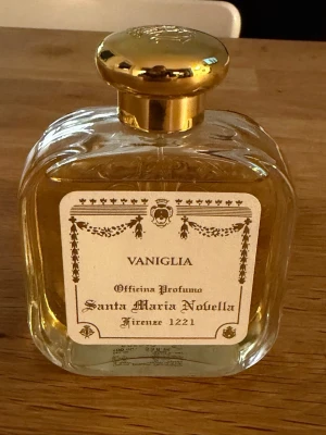 Santa Maria Novella Vaniglia parfym - Exklusiv parfymflaska från Santa Maria Novella med doften Vaniglia. Flaskan är genomskinlig med en elegant guldig kork och klassisk etikett i vitt och guld. Perfekt för dig som gillar lyxiga och tidlösa dofter med vaniljtoner.