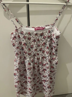 Blommig vit linnetopp  - Supersöt linnetopp med smala axelband och smock upptill. Toppen är vit med små blommor i rött och blått och tunna ränder i guld🤩, den är i storlek 158/164 men passar även som ”S”