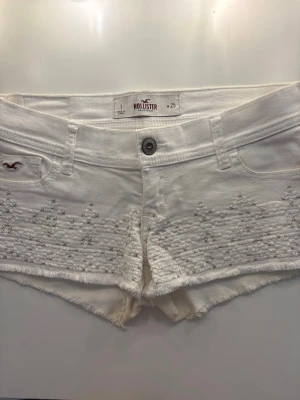 Hollister jeansshorts  - Snygga vita shorts från Hollister med broderade detaljer framtill och råa kanter. Klassisk femficksmodell med knapp och dragkedja. Perfekta för sommaren och ger en fräsch och avslappnad look.