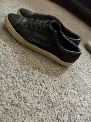 Svarta Saint Laurent sneakers - Svarta sneakers från Saint Laurent med vit sula och handskriven logga på sidan. Skorna har klassisk låg profil, rund tå och svarta skosnören. Tillverkade i slätt läder för en clean och stilren look. Skriv om ni har några frågor 
