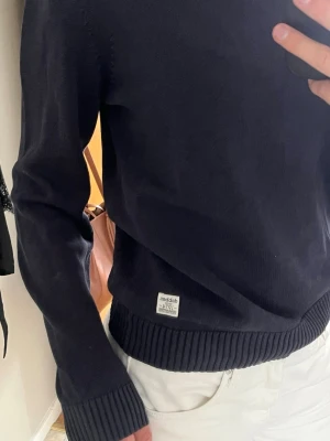 Mörkblå stickad tröja - Säljer en enkel och stilren mörkblå stickad tröja med rund hals och ribbade muddar vid ärmslut och nederkant. Tröjan har en liten vit etikett nertill och är i bomull.❣️