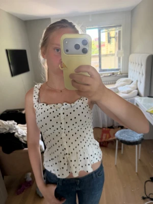 Vit prickig linnetopp med volang - Supersöt vit linnetopp med svarta prickar och volangkant. Toppen har breda axelband, knappar framtill och smockad midja som ger en snygg passform. Perfekt till jeans eller kjol för en trendig look.