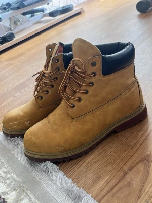 Beige snörade stövletter i mocka-look - Snygga beige stövletter med snörning och kraftig sula. Skorna har en vadderad svart kant upptill och är gjorda i ett material som liknar mocka. Perfekta för dig som gillar en robust och trendig stil.