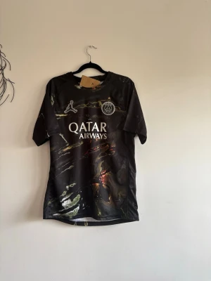 Svart PSG matchtröja med tryck - Säljer en svart PSG matchtröja med marmorerat mönster och tryck på bröstet samt ryggen. Framtill står det 'Qatar Airways' och PSG-loggan, på ryggen står det 'D. Doué' och nummer 14. Tröjan har korta ärmar och sportig passform.(kan gå ner lite i pris vid snabb affär)