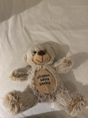 Mjuk nallebjörn med text - Söt mjuk nallebjörn i ljusbrunt fluffigt tyg med broderad nos och ett stort leende. På magen står det 'Världens bästa älskling' broderat i svart. Perfekt som present till någon du tycker om.
