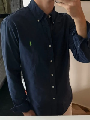 Mörkblå skjorta från Ralph Lauren - Klassisk mörkblå skjorta från Ralph Lauren med button-down krage och grönt broderat logotyp på bröstet. Skjortan har långa ärmar, vita knappar och är tillverkad i mjuk bomull. Perfekt för en stilren och avslappnad look. Ganska urtvättad, därav priset 