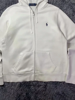Vit hoodie från Polo  - Snygg vit hoodie från Polo Ralph Lauren med dragkedja framtill, huva med dragsko och klassisk blå logga broderad på bröstet. Hoodien har känguruficka och är gjord i mjukt material som känns riktigt skönt mot huden. Perfekt för en clean och stilren look.