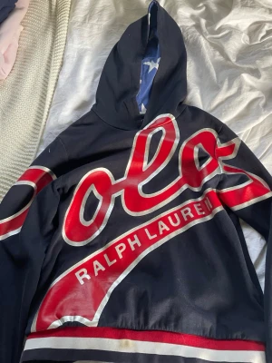 Mörkblå hoodie från Ralph Lauren - Snygg mörkblå hoodie från Ralph Lauren med stor röd och vit POLO-logga över bröstet och texten 'Ralph Lauren' i vitt. Hoodien har huva med blått innerfoder och vita stjärnor. Perfekt för dig som gillar streetwear och vill sticka ut.