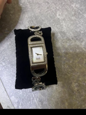 Silverfärgad D&G klocka med stenar - Snygg och unik klocka från D&G i silverfärg med rektangulär urtavla och gnistrande stenar runt ramen. Armbandet har stora D&G-bokstäver som länk och pärlemorskimrande urtavla. Perfekt accessoar för dig som gillar statement-smycken.