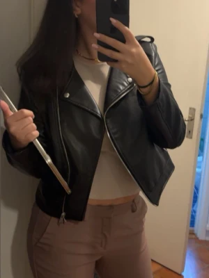 Svart kort skinnjacka - Snygg svart skinnjacka i kort modell med klassisk bikerskärning och silvriga detaljer. Jackan har breda slag, dragkedja och en cool, stilren look som passar perfekt till många outfits.