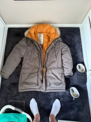 Brun parkas med fuskpäls och orange foder - Snygg brun parkas med varm orange foder och huva med fuskpäls. Jackan har stora fickor framtill, dragkedja och tryckknappar. Perfekt för kalla dagar och har en klassisk, rymlig passform.