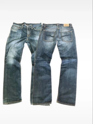 Nudie jeans slitningar - Säljer ett par blå jeans med snygga slitningar framtill. Jeansen har klassisk femficksdesign och kontrastsömmar. Materialet är denim i bomull och passformen är avslappnad. Perfekta för en casual look.