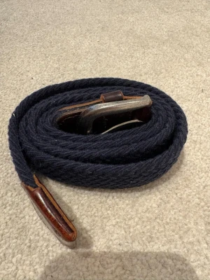 Mörkblått flätat bälte med läderdetaljer - Snyggt mörkblått flätat bälte med bruna läderdetaljer och metallspänne. Perfekt för att ge din outfit en avslappnad men ändå stilren touch. Passar till jeans eller chinos och är lätt att justera efter önskad längd.stretch 