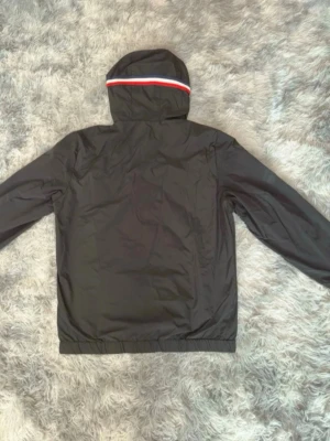 Moncler windbreaker  - Svart vindjacka från Moncler med huva och röd-vit-blå rand på huvan. Jackan har resår i midjan och ärmarna samt Moncler-logga på vänster ärm. Tillverkad i ett lätt, vattenavvisande material som passar perfekt för blåsiga dagar.