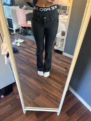 Svarta bootcut jeans med fickor - Säljer ett par svarta bootcut jeans med klassisk femficksdesign och silverfärgade knappar på bakfickorna. Jeansen har normal midja och lätt utsvängda ben. Materialet är stretchigt denim som sitter snyggt och bekvämt.