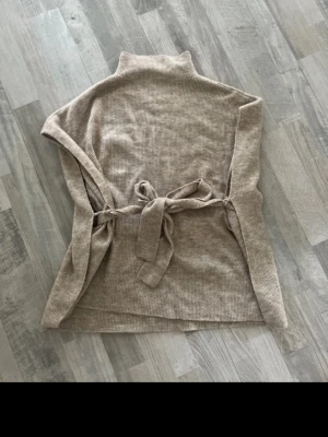 Beige ribbad polotröja från Kappahl - Säljer en beige ribbad polotröja från Kappahl med knytband i midjan. Tröjan har hög krage och är ärmlös, vilket gör den perfekt för lager på lager. Materialet är mjukt och stretchigt för en bekväm passform.