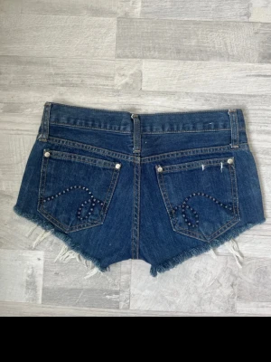 Blå jeansshorts med slitningar och nitar - Säljer ett par blå jeansshorts med råa, fransiga kanter och slitningar framtill. Baksidan har coola nitar i mönster på fickorna. Klassisk femficksmodell med knapp och dragkedja. Perfekta för sommaren och ger en avslappnad streetstyle-vibe.