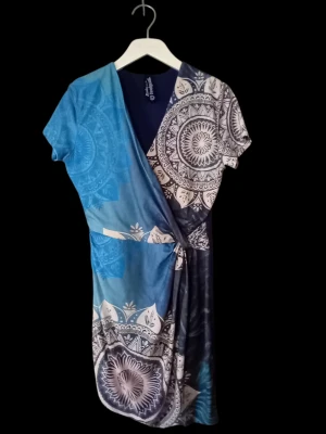 Blå klänning med omlott överdel från Desigual L - Unik kort omlottklänning från Desigual i blå och vita toner med mandalainspirerat mönster. Klänningen har korta ärmar och knyt i midjan för en snygg passform. Perfekt för dig som gillar färg och mönster! Mjuk och följsam.