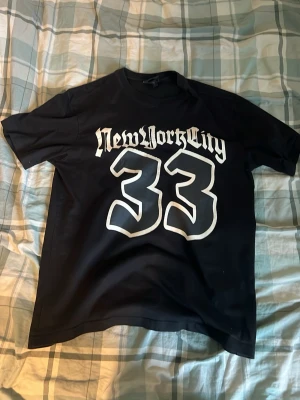 Svart t-shirt med New York City-tryck - Cool svart t-shirt med stort vitt tryck där det står 'New York City' och siffran 33 på framsidan. Klassisk passform och rund halsringning, perfekt för en streetwear-look.