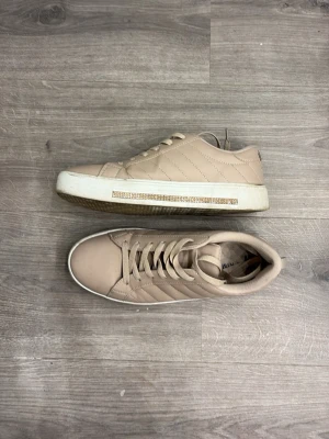 Beige sneakers med strassdetalj - Snygga beige sneakers i konstläder med vita sulor och strassdetaljer på sidan. Skorna har klassisk låg modell, ton-i-ton snörning och diskreta sömmar som ger en stilren look. Perfekta för dig som vill ha något neutralt men ändå med lite bling.